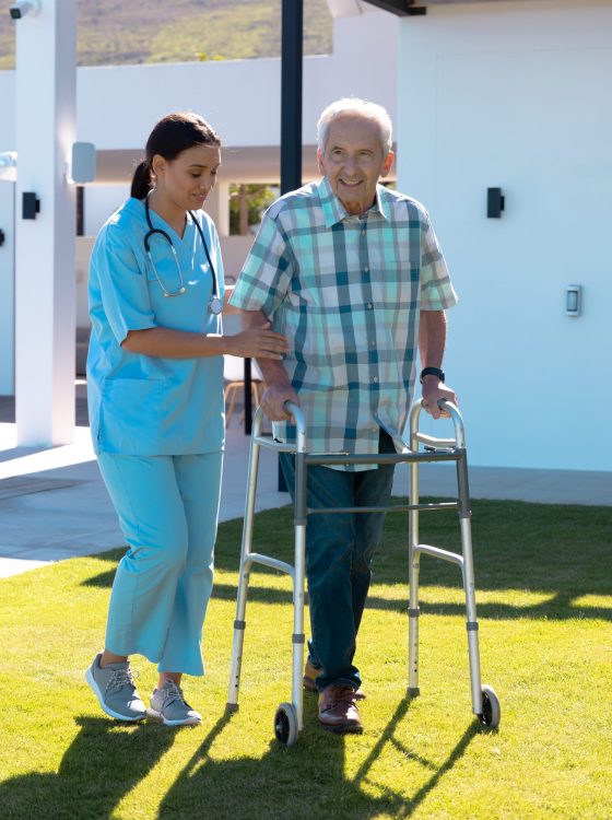 Biracial female doctor assisting smiling caucasian senior man in walking with walker in yard