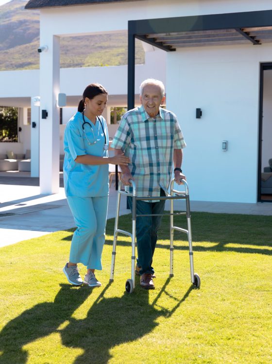Biracial female doctor assisting smiling caucasian senior man in walking with walker in yard. House, summer, unaltered, healthcare, patient, retirement, support, recovery and disability concept.