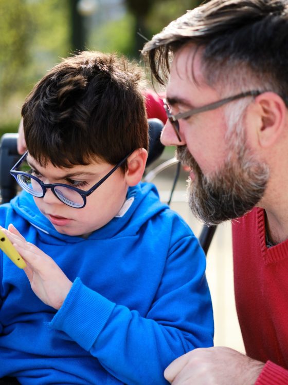 Disabled child on wheelchair watching something on the mobile phone while enjoying outdoors with his father. Technology and disability people concept.