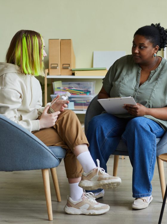 African female social worker talking to difficult teenage girl while they sitting on armchairs at office