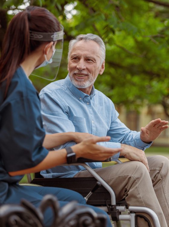Talkative aged man, recovering patient in wheelchair having conversation with his nurse in protective face shield, resting together in the park near hospital