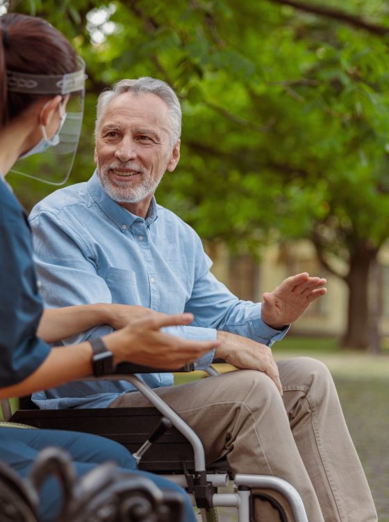 Talkative aged man, recovering patient in wheelchair having conversation with his nurse in protective face shield, resting together in the park near hospital. Support, rehabilitation concept